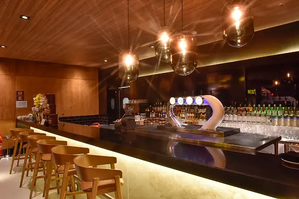 Balcão do bar americano com banquetas de madeira, luminárias de vidro e prateleira de bebidas