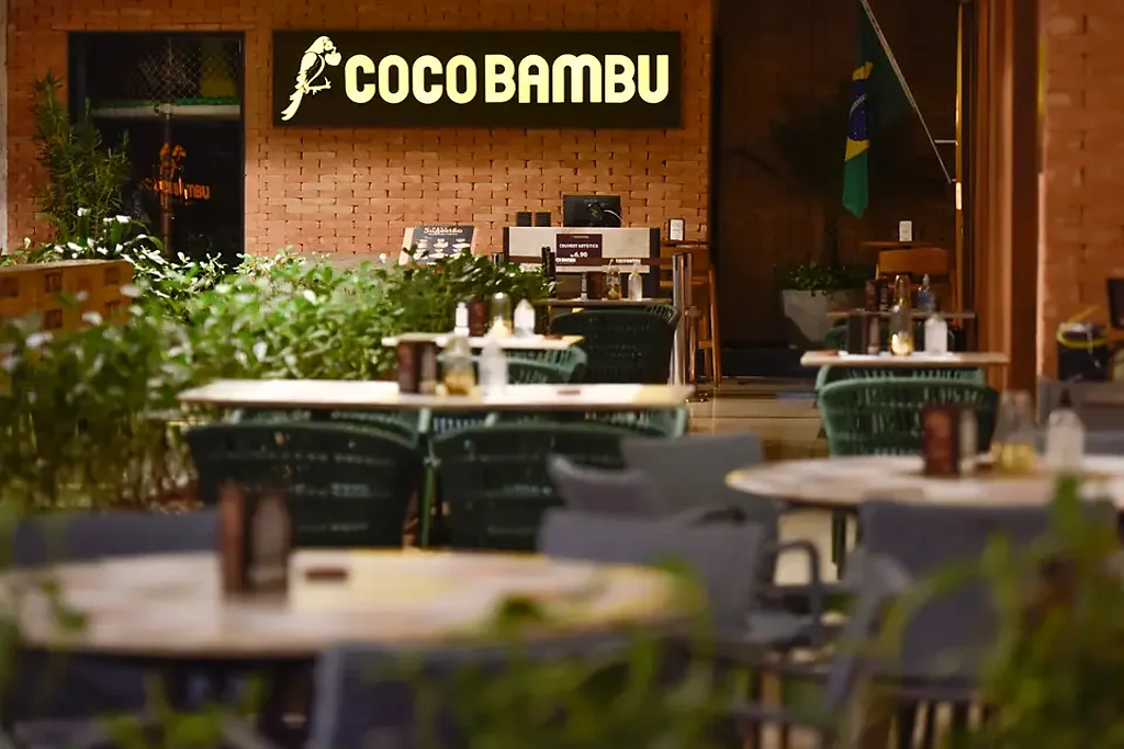 Entrada do Coco Bambu Botafogo com letreiro iluminado sobre a parede de tijolos e mesas na varanda