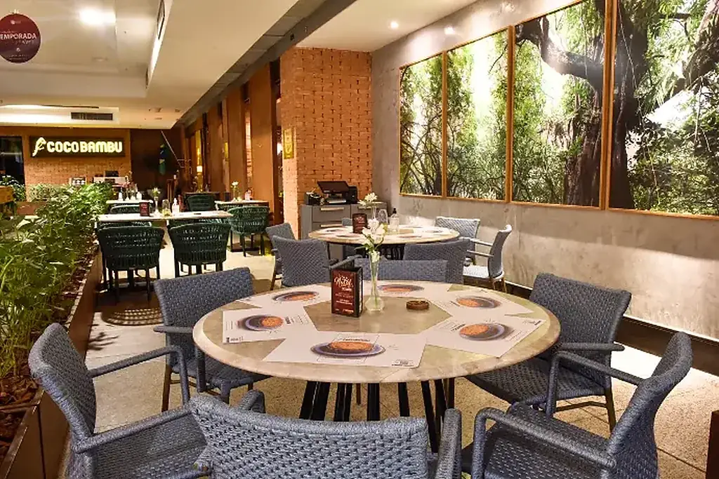 Varanda do restaurante com mesas redondas, cadeiras de fibra e painéis de vegetação nas janelas